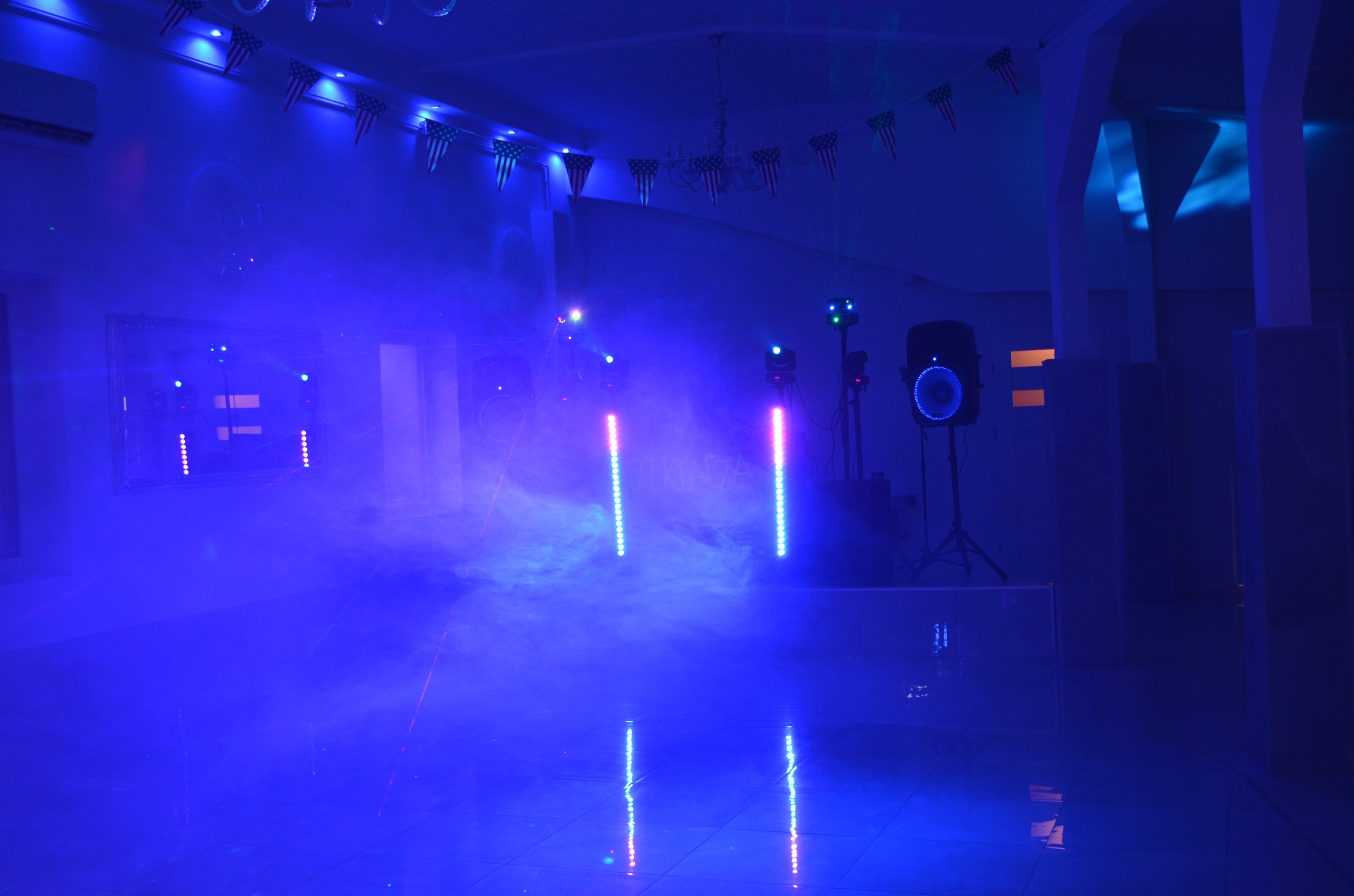 Oświetlenie imprezowe w pomieszczeniu z klimatyzacją: niebieskie światła, kolumny LED, głośniki i dekoracje flagami USA. Klimatyzator widoczny na ścianie.