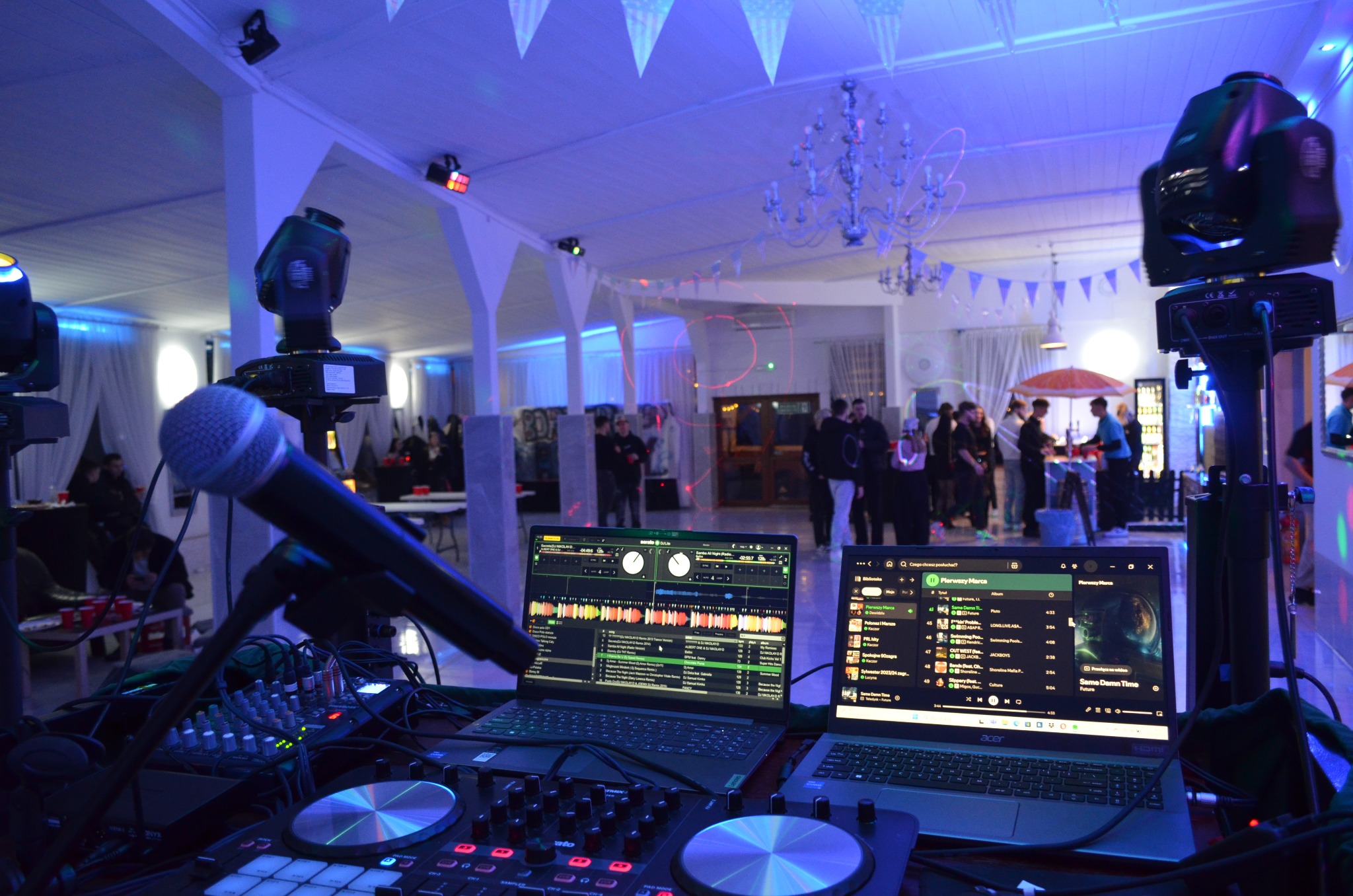 Sprzęt DJ-ski z dwoma laptopami i mikrofonem na tle sali z oświetleniem imprezowym i bawiącymi się ludźmi. Widoczne dekoracje i żyrandol.