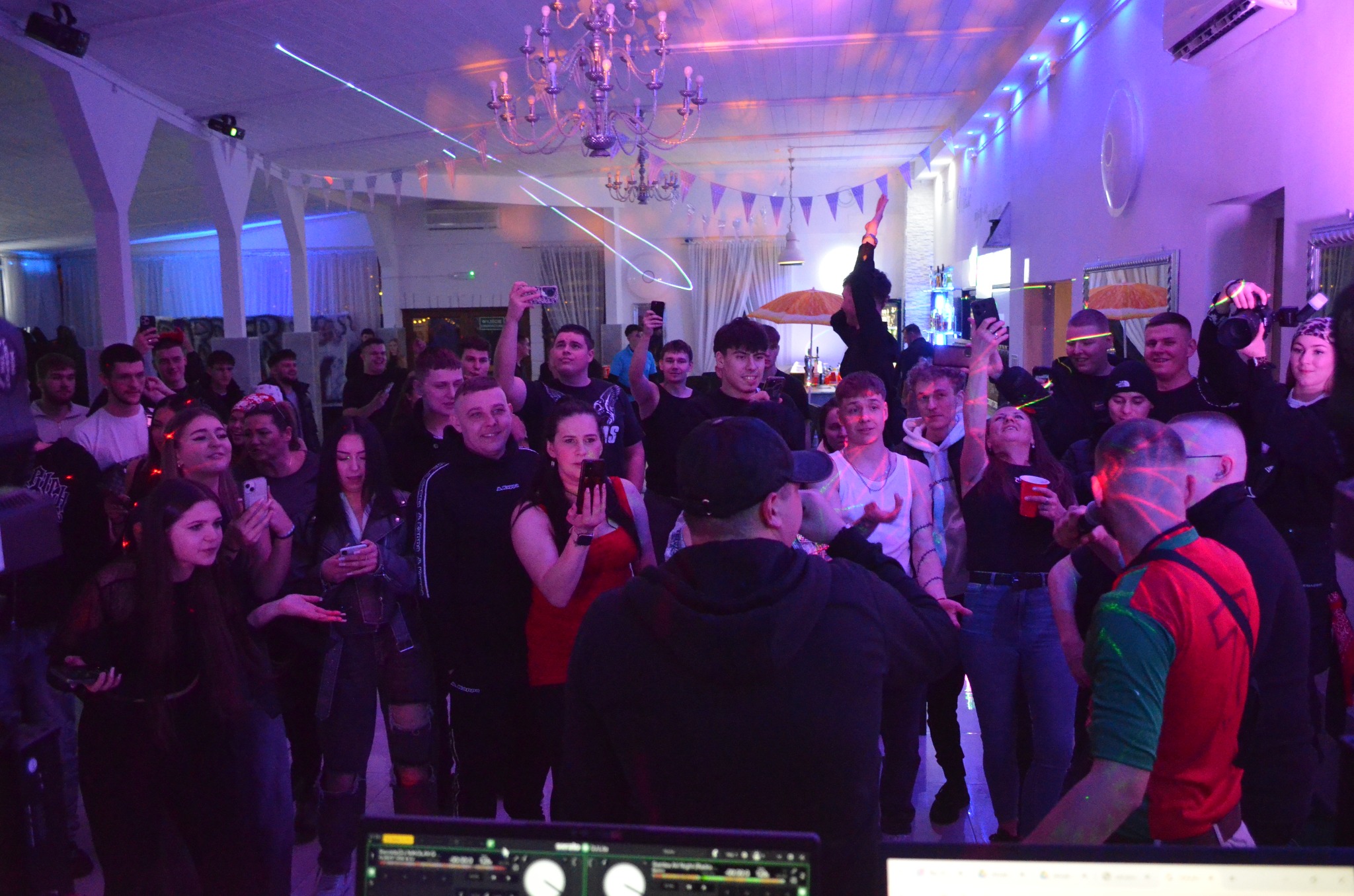 Impreza w lokalu z klimatyzacją: tłum ludzi, DJ, oświetlenie dyskotekowe i wiszący klimatyzator na ścianie. Laserowe efekty świetlne.