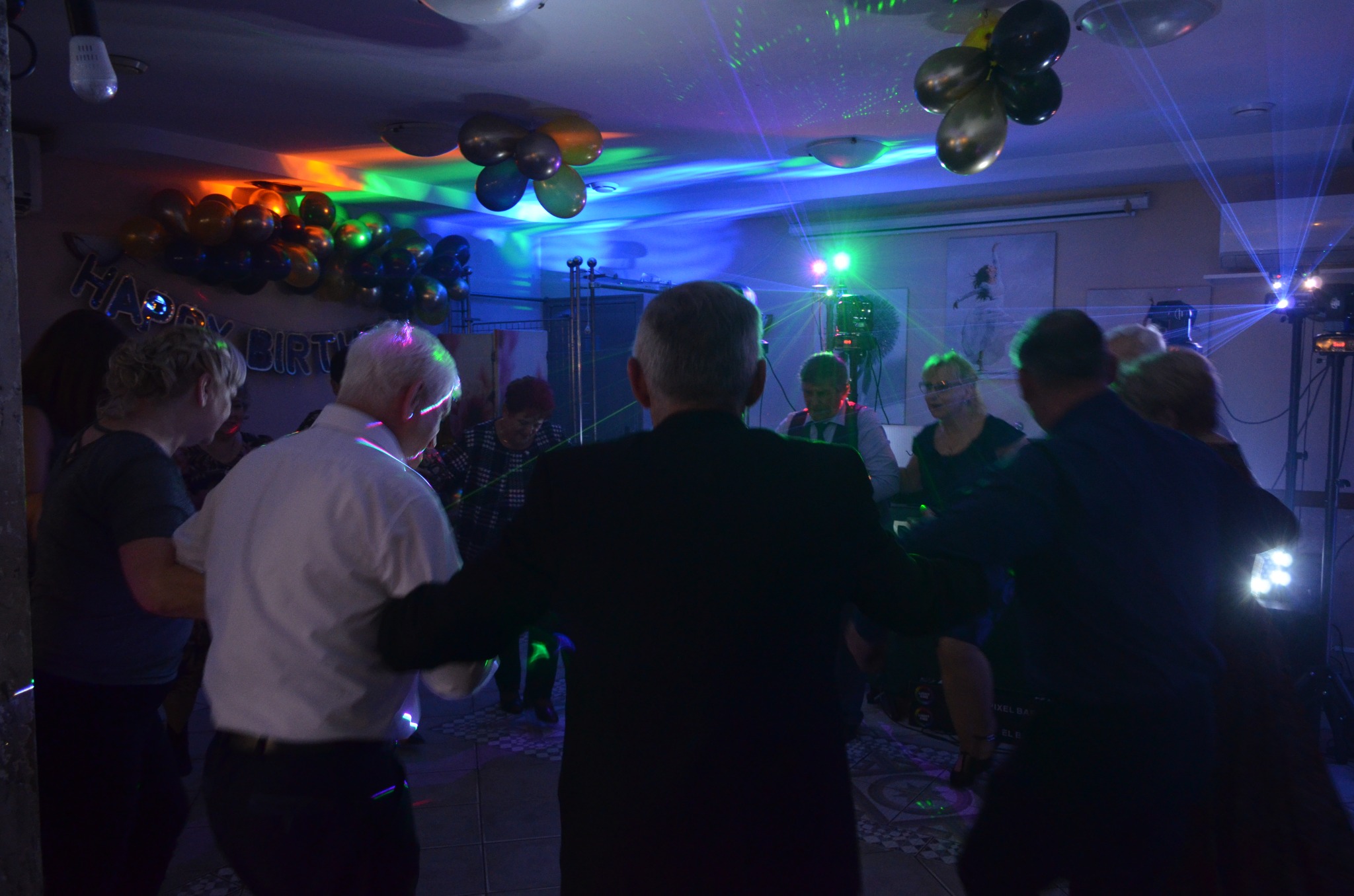 Zabawa taneczna w pomieszczeniu z dekoracjami urodzinowymi, oświetlona kolorowymi światłami dyskotekowymi i balonami pod sufitem.