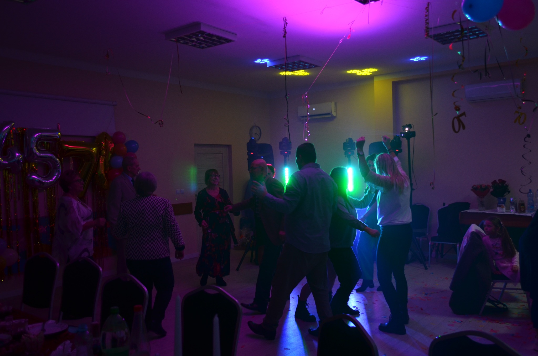 Impreza w pomieszczeniu z klimatyzacją ścienną, oświetlona kolorowymi światłami. Tańczący ludzie, balony i dekoracje urodzinowe.