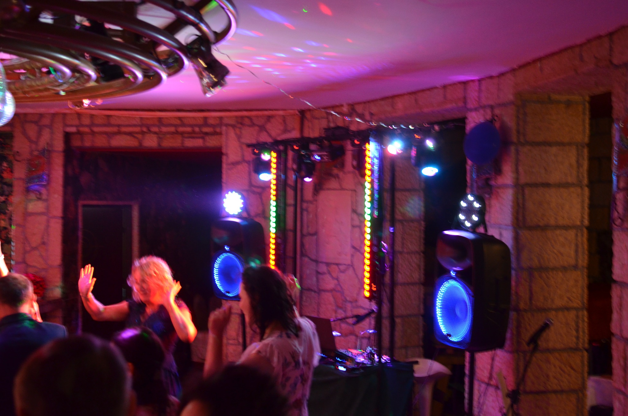 Impreza w lokalu z oświetleniem dyskotekowym i nagłośnieniem. Parkiet wypełniony tańczącymi osobami, kolorowe światła i głośniki tworzą klimat zabawy.