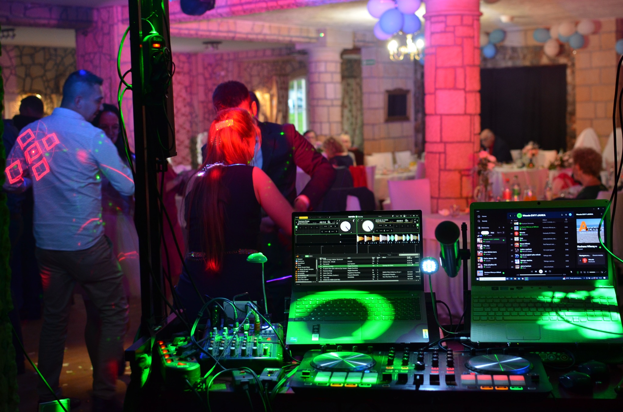 DJ na weselu: stanowisko z mikserem, laptopami i oświetleniem w zielonych barwach, w tle tańczący goście w sali z kolumnami i dekoracjami.