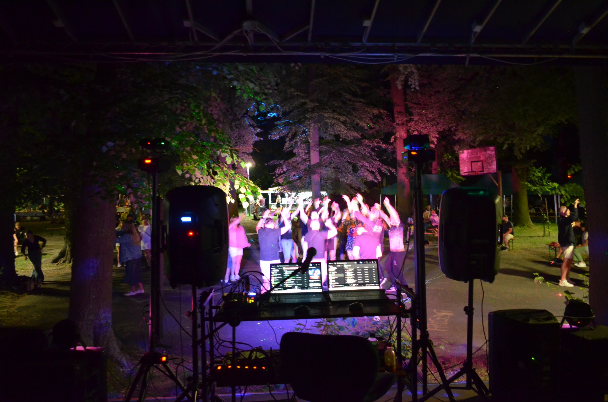 Impreza plenerowa z DJ-em i nagłośnieniem. Dwa laptopy na stole, głośniki po bokach, w tle bawiący się ludzie podświetleni kolorowymi światłami.