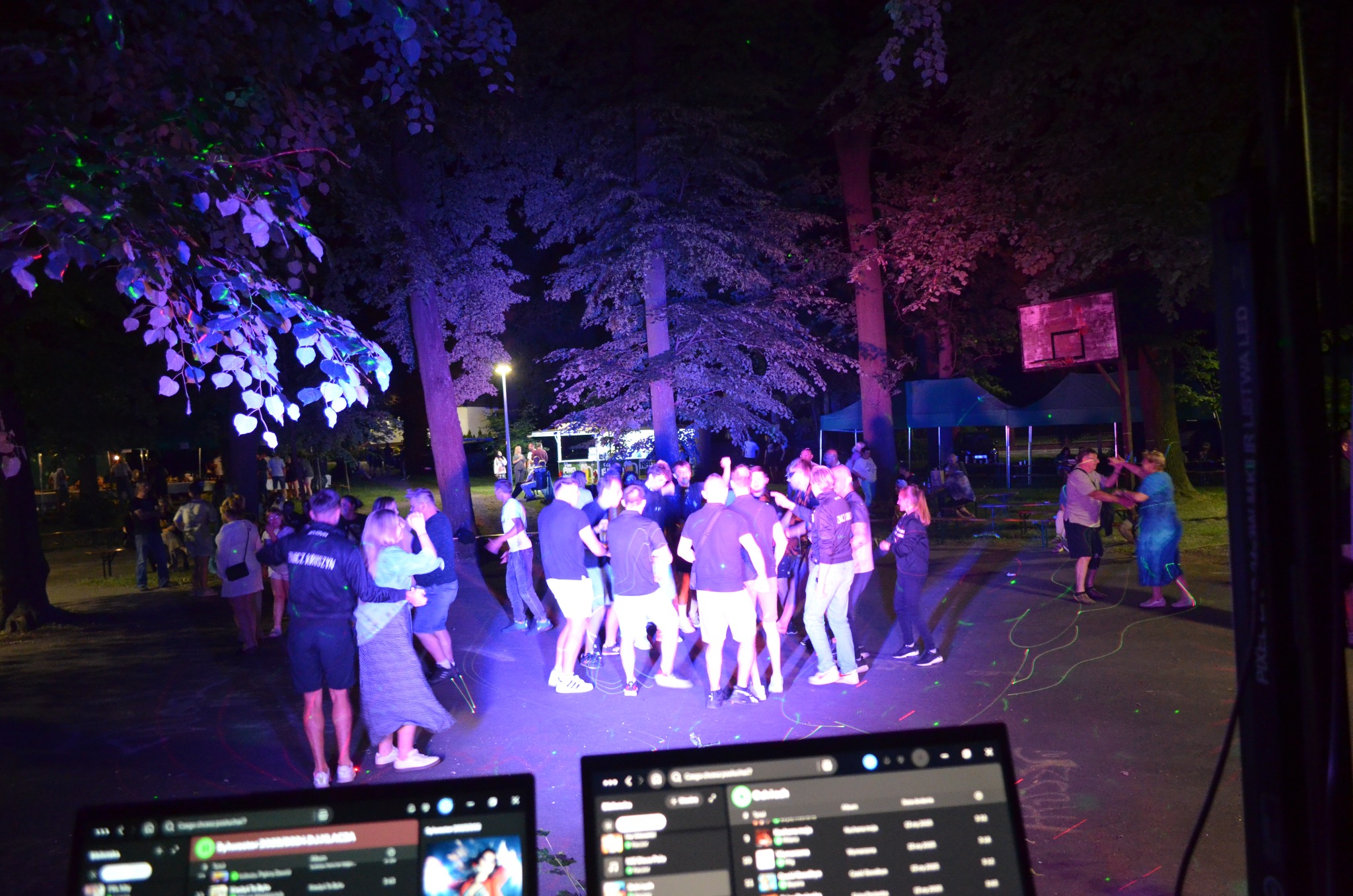 Impreza plenerowa nocą: tańczący ludzie pod drzewami oświetlonymi kolorowymi światłami, widoczne ekrany laptopów DJ-a. Atmosfera zabawy i muzyki.