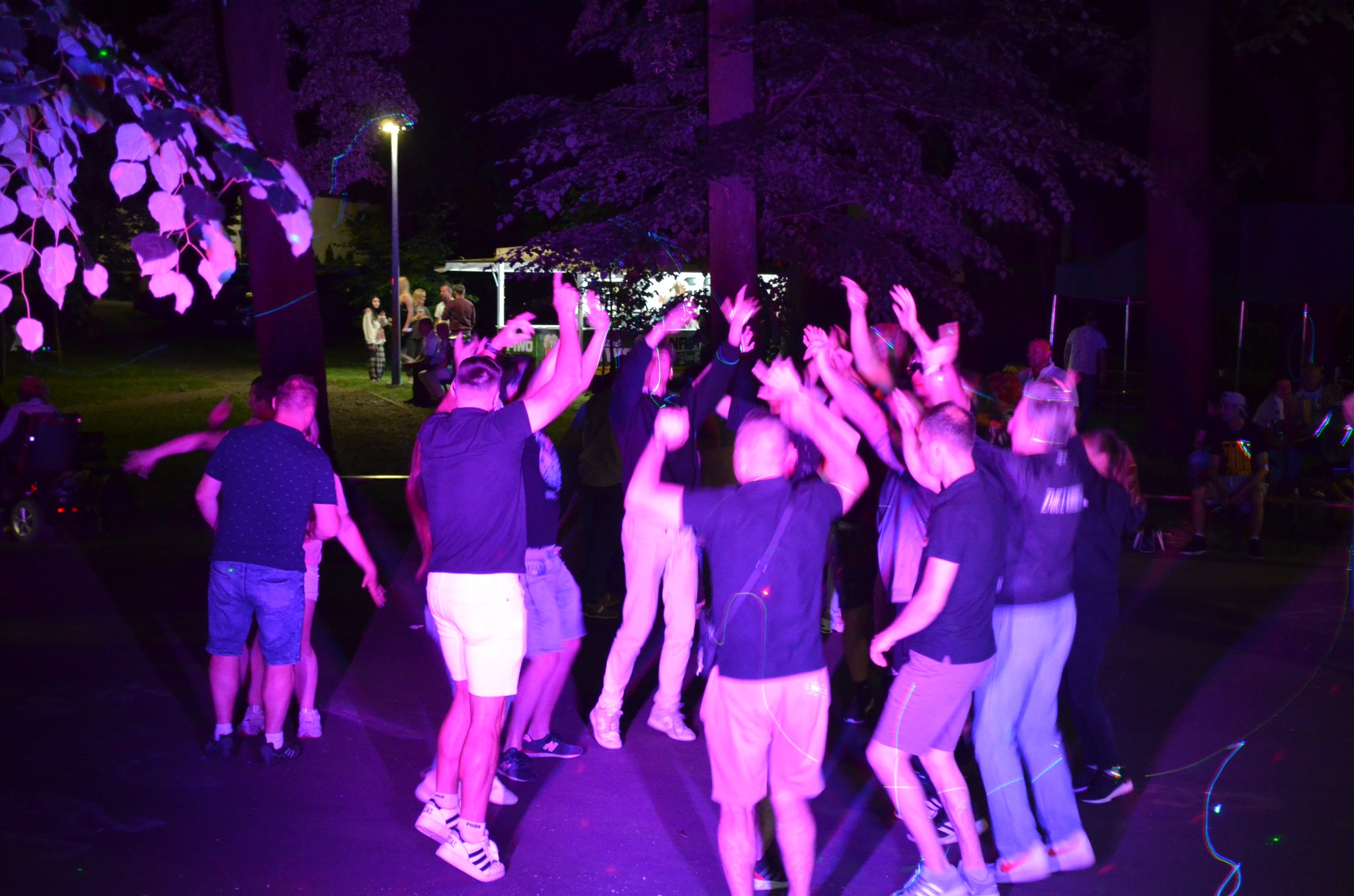 Rozbawiona grupa ludzi tańczy w fioletowym oświetleniu na zewnątrz, prawdopodobnie podczas imprezy plenerowej. W tle widoczne drzewa i budka.