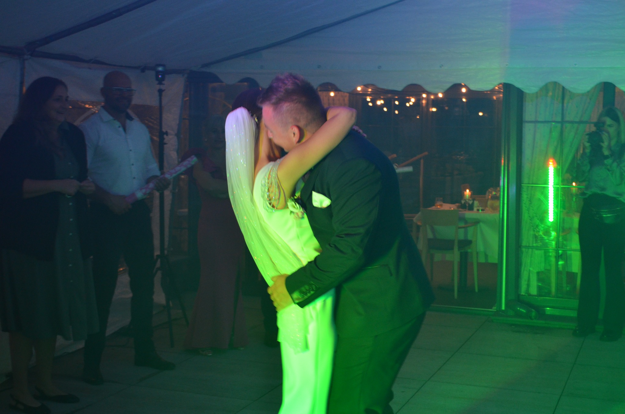 Para młoda w tańcu podczas wesela w namiocie, oświetlonym zielonymi i niebieskimi światłami. W tle dekoracje i goście.