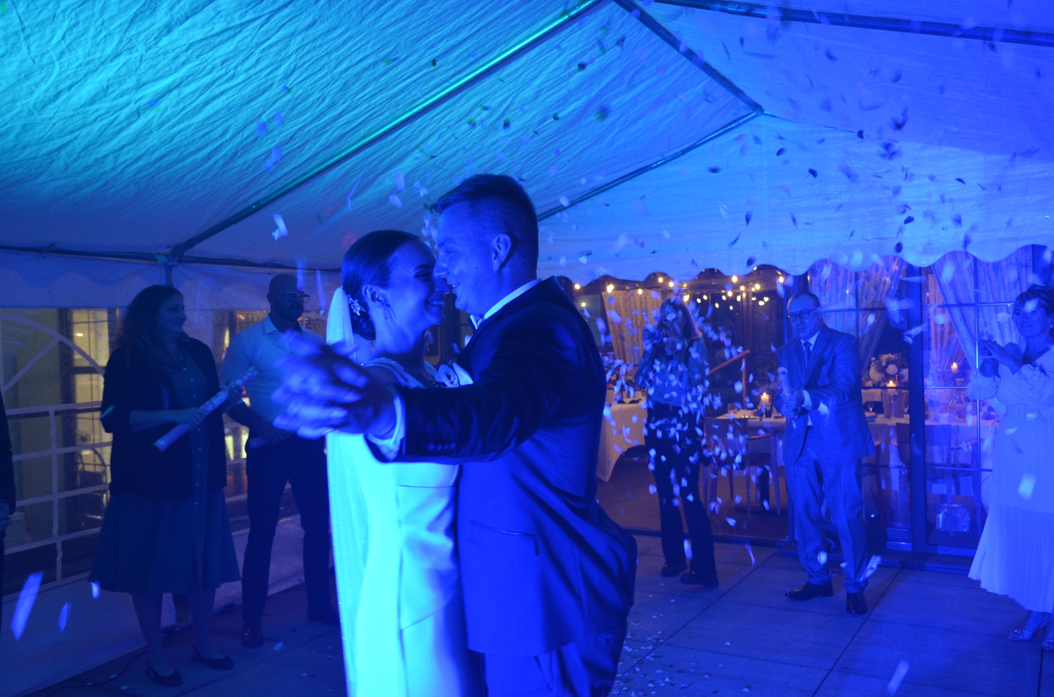 Para młoda tańczy pod namiotem oświetlonym na niebiesko, z konfetti w powietrzu. W tle goście i dekoracje.