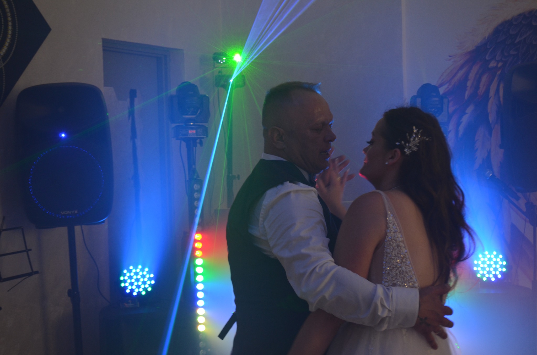 Para tańczy w objęciach, oświetlona laserami i reflektorami. Widoczny sprzęt nagłośnieniowy i oświetleniowy. Dekoracja ściany z motywem piór.