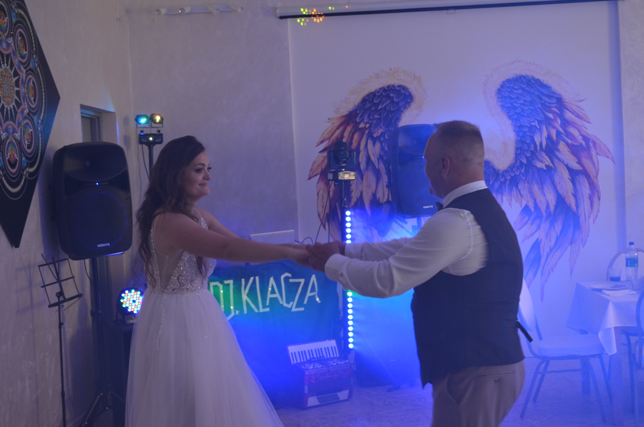 Para tańczy na weselu przy dekoracji ze skrzydłami, oświetleniu DJ i akordeonie. DJ KlaCza zapewnia oprawę muzyczną.
