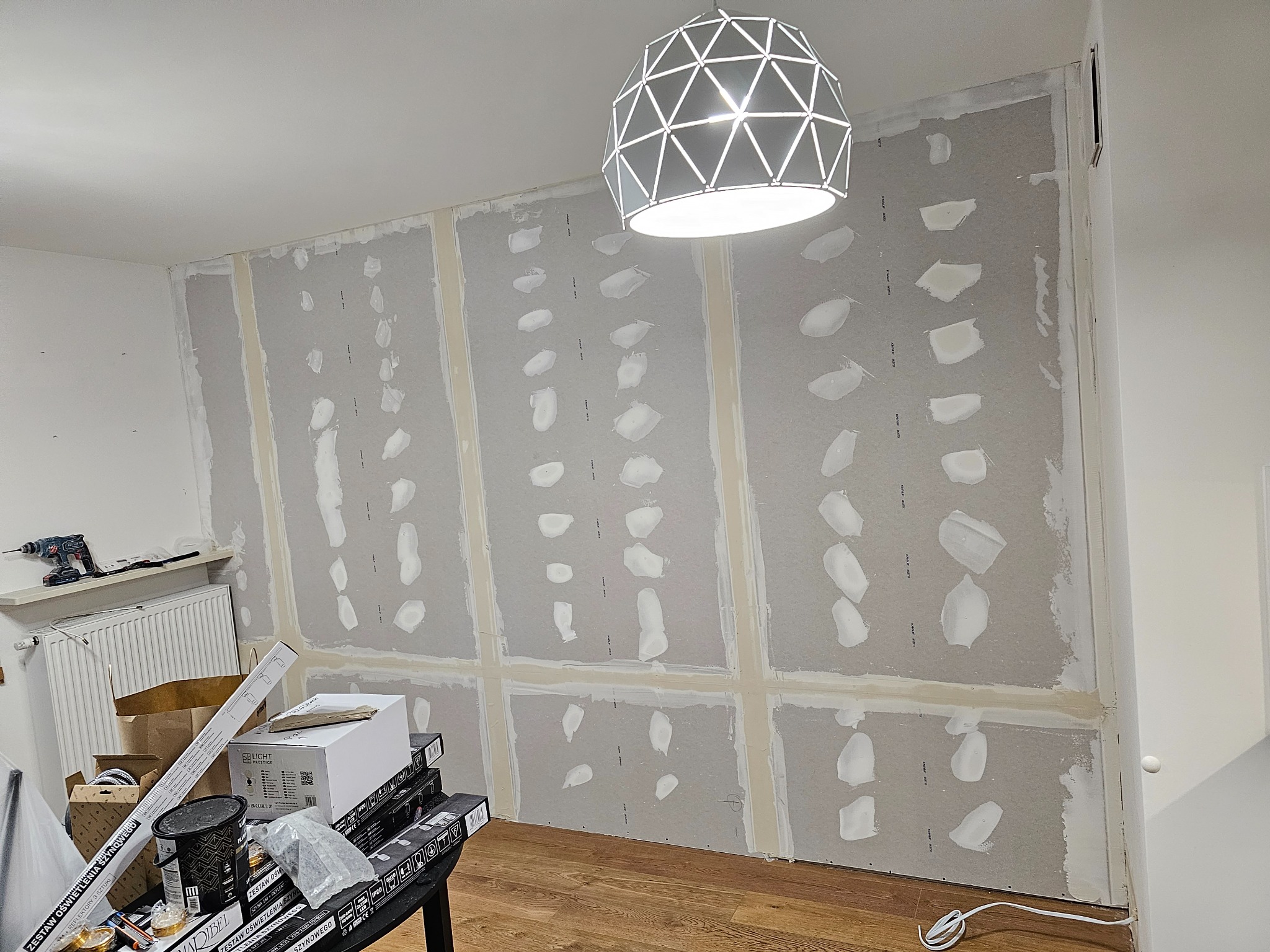 Ściana z płyt kartonowo-gipsowych w trakcie szpachlowania, widoczne łączenia płyt i nałożona gładź, w tle lampa sufitowa i narzędzia na stole.
