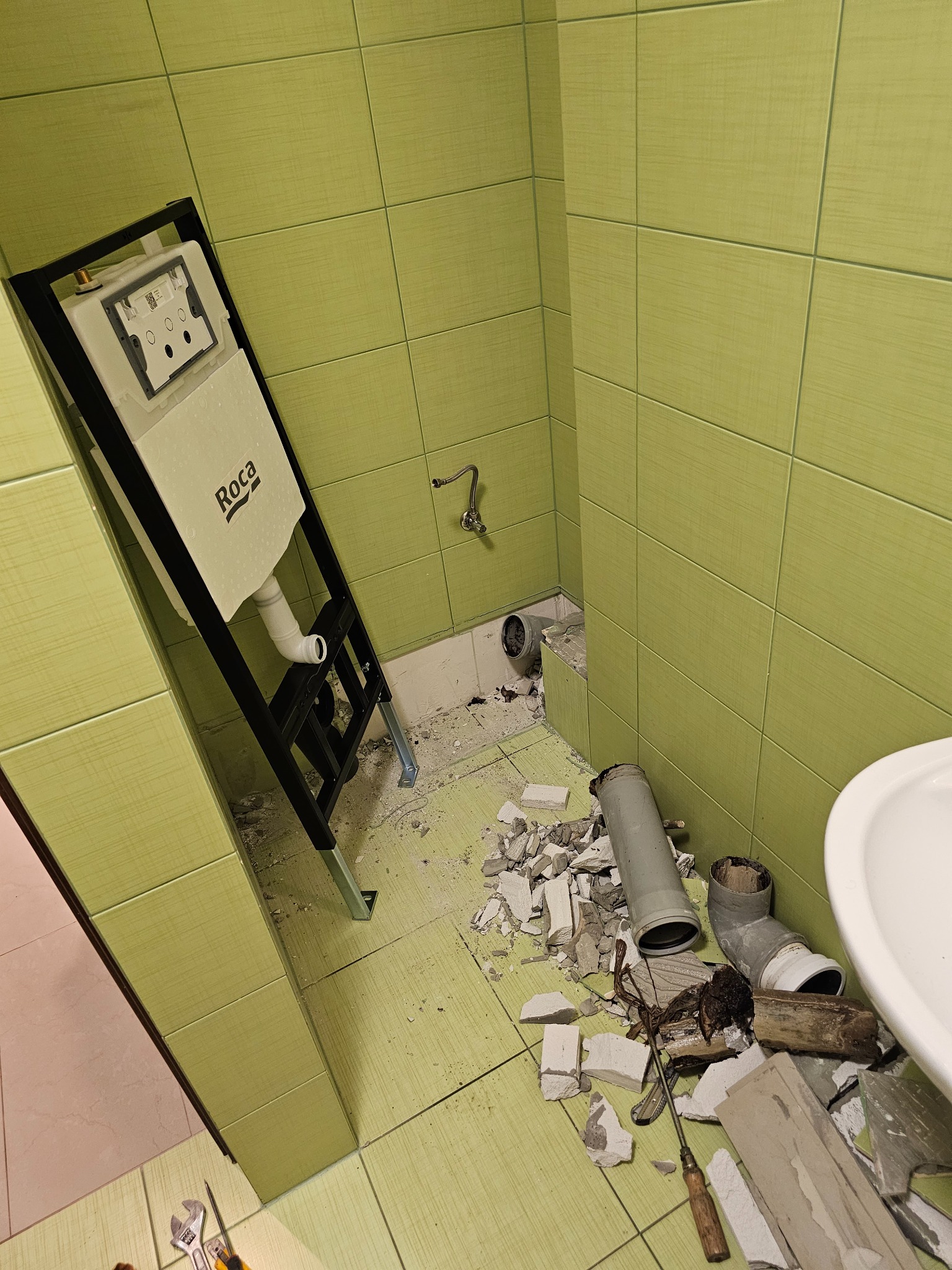 Wnętrze łazienki w trakcie remontu: zamontowany stelaż podtynkowy Roca, widoczne fragmenty potłuczonych płytek, rury kanalizacyjne i narzędzia.