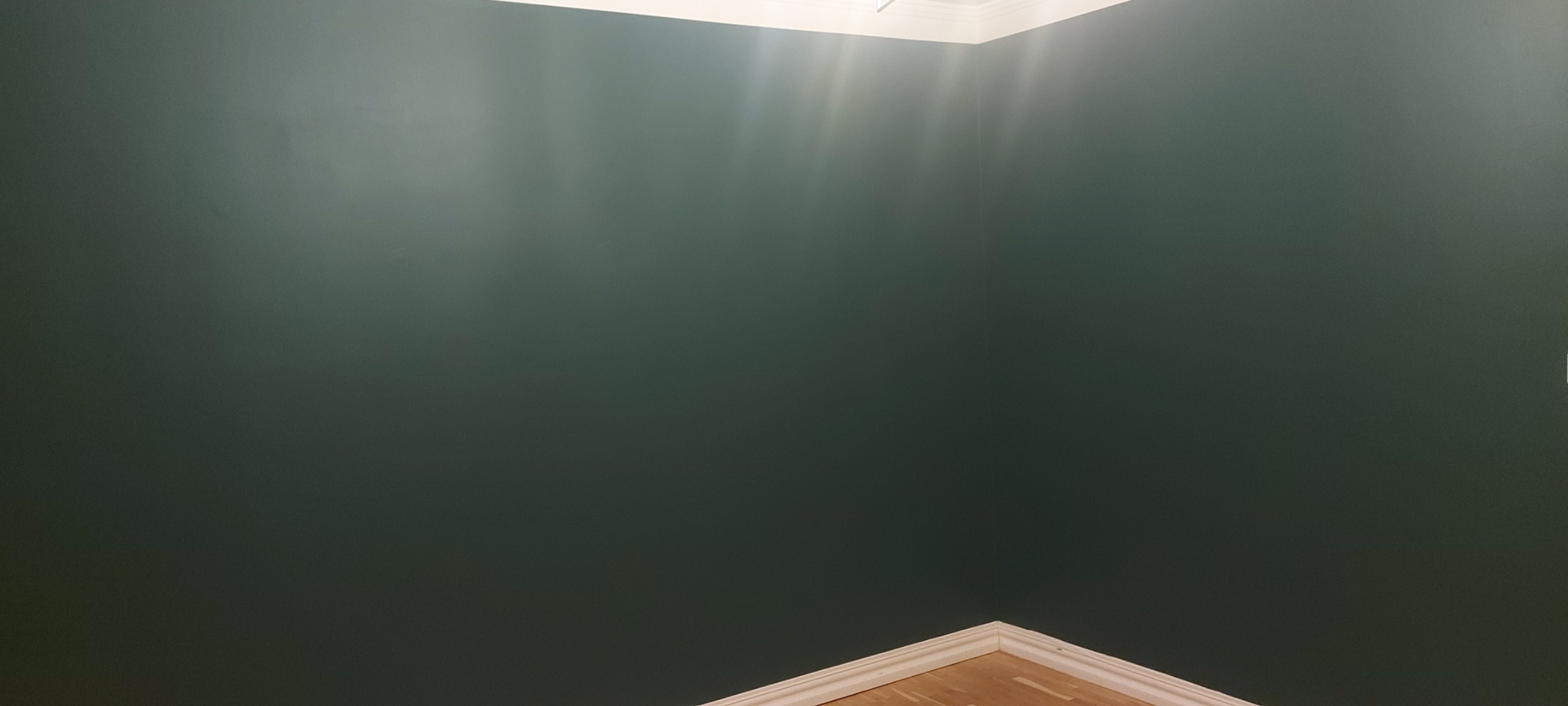 Ściana pomalowana na ciemnozielony kolor z widocznymi refleksami światła, biały sufitowy gzyms i fragment drewnianej podłogi z listwą przypodłogową.