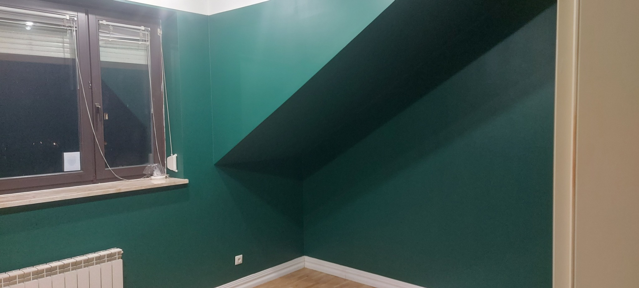 Pomieszczenie po odświeżeniu ścian farbą w odcieniach zieleni, widoczny fragment okna z żaluzjami, grzejnik oraz wykończona podłoga.