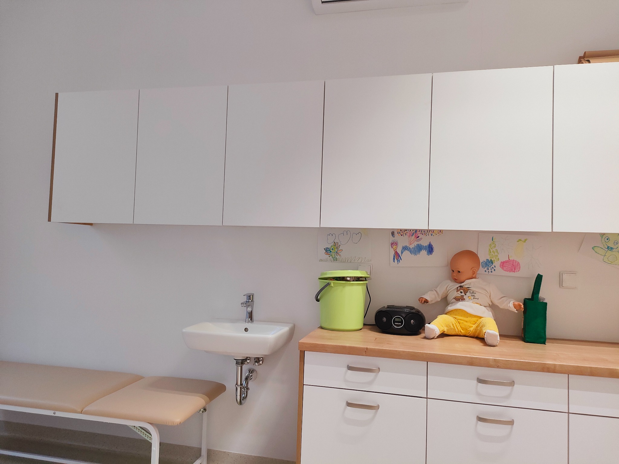 Jasny gabinet z umywalką, kozetką, szafkami i lalką na blacie, ozdobiony dziecięcymi rysunkami na ścianie.