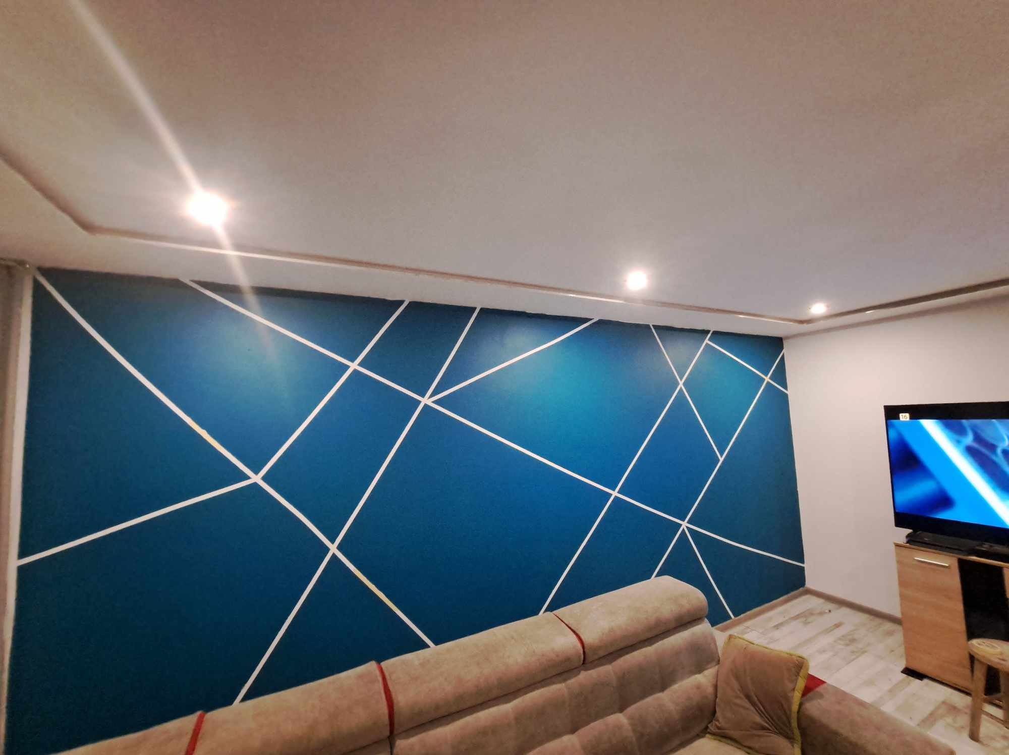 Ściana pomalowana na niebiesko z białymi geometrycznymi wzorami, widoczny fragment sofy i telewizor w rogu pokoju.