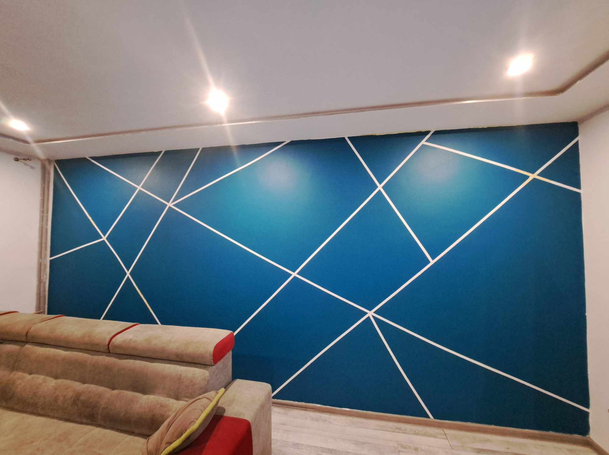Ściana pomalowana na niebiesko z geometrycznym wzorem z białych linii, widoczny fragment beżowej sofy z czerwonymi akcentami, oświetlenie sufitowe.