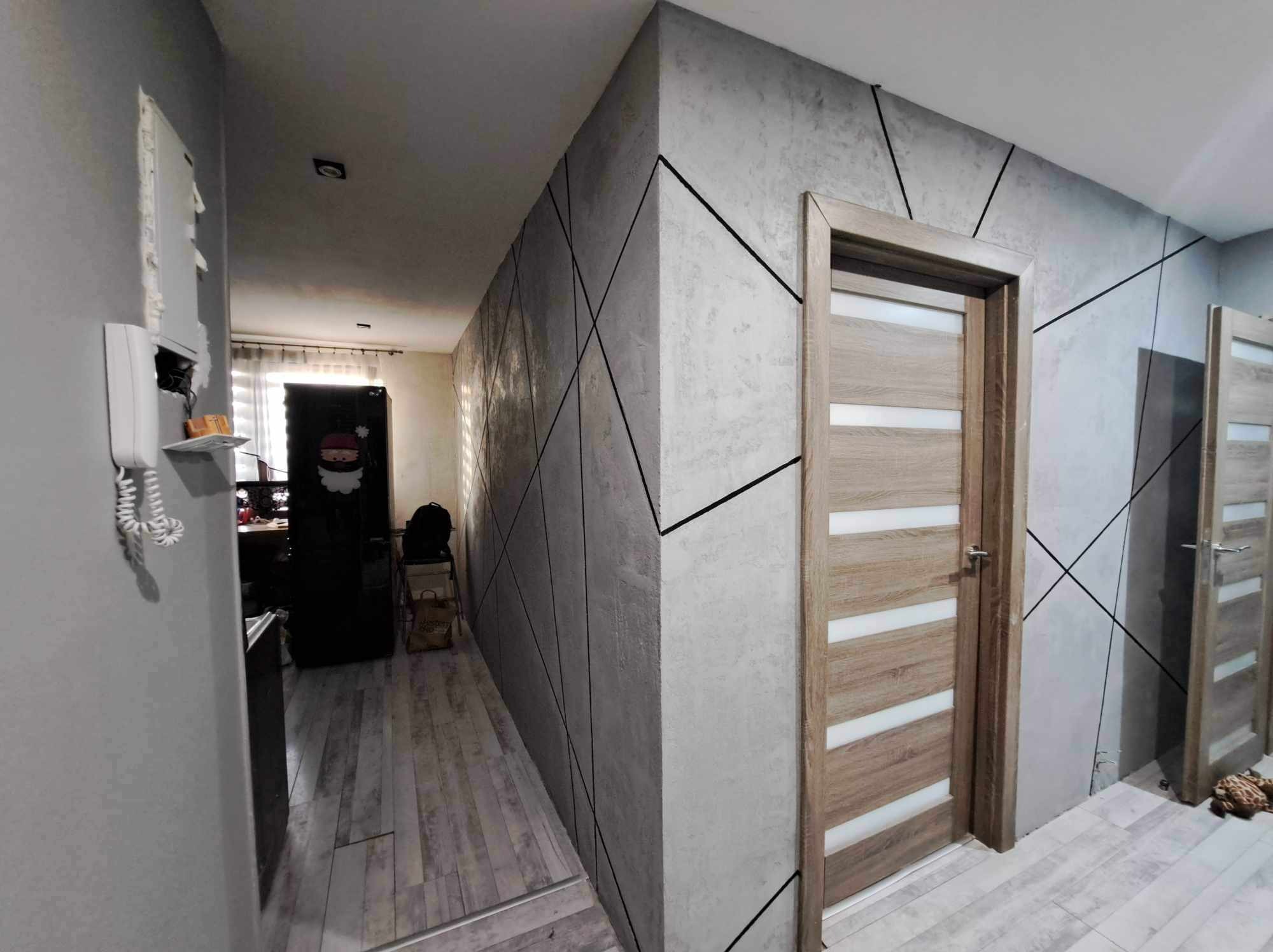 Korytarz z szarą ścianą o fakturze betonu, zdobioną czarnymi, ukośnymi liniami, oraz dwiema drewnianymi drzwiami z białymi, poziomymi przeszkleniami.