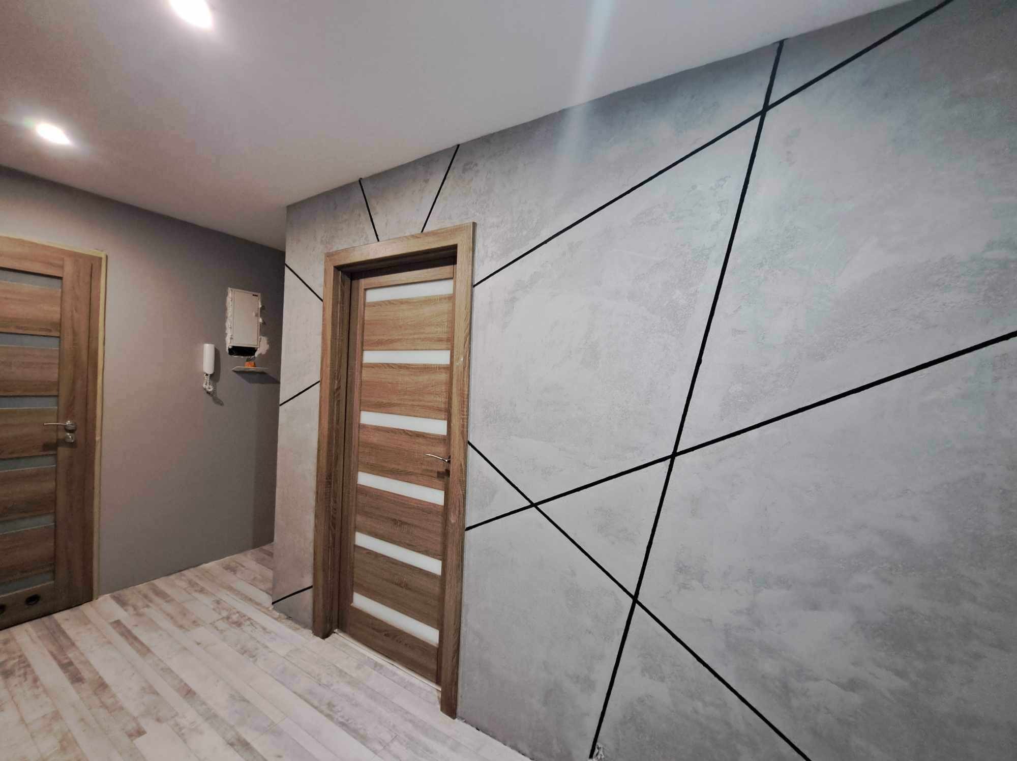 Korytarz po remoncie z szarą ścianą o fakturze betonu, dekorowaną czarnymi liniami tworzącymi geometryczny wzór, drewniane drzwi z białymi panelami i podłoga imitująca drewno.