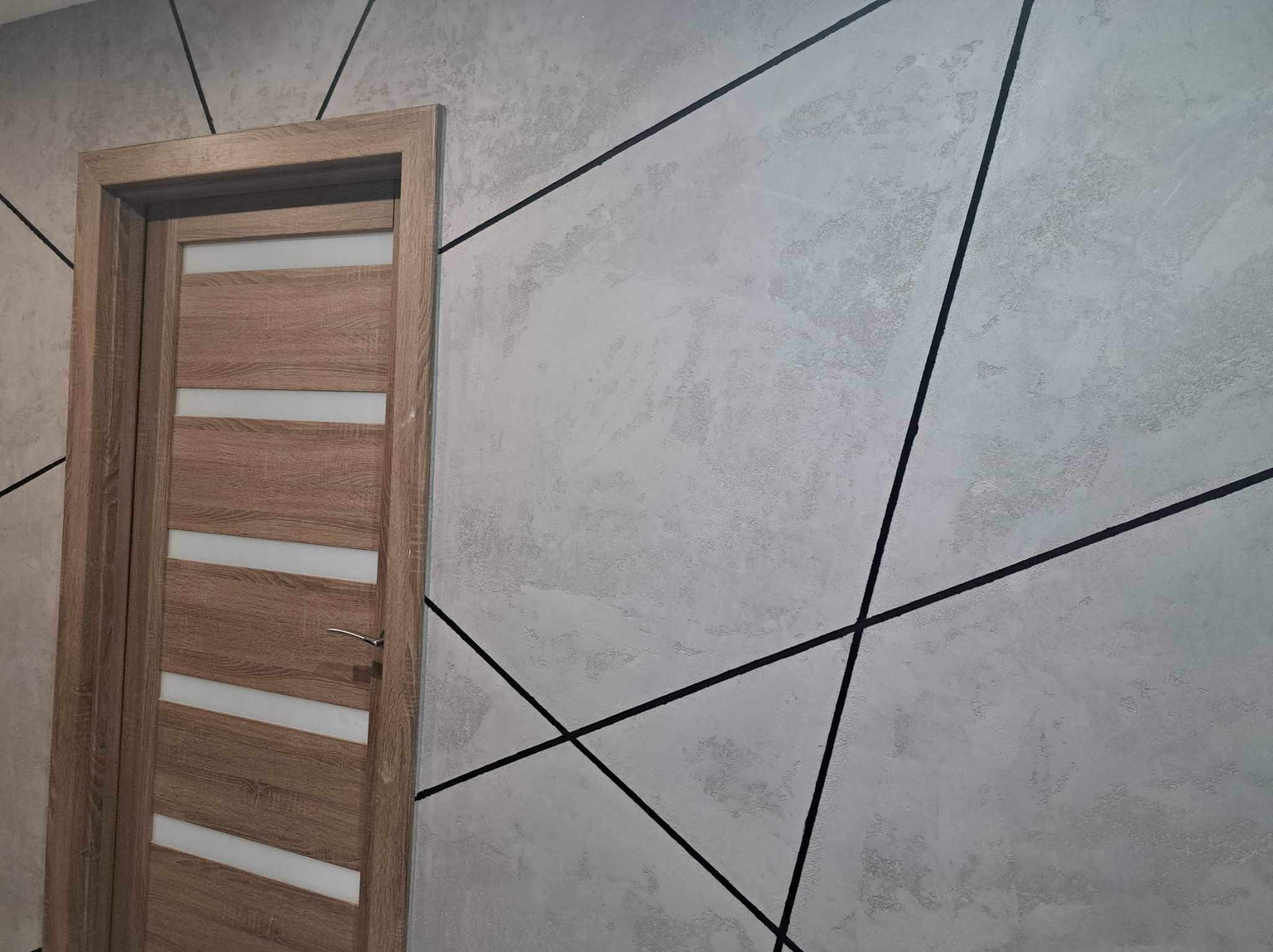 Nowoczesne wykończenie ściany w Zawierciu z szarą strukturą tynku i geometrycznym wzorem z czarnych linii, obok drewniane drzwi z przeszkleniami.