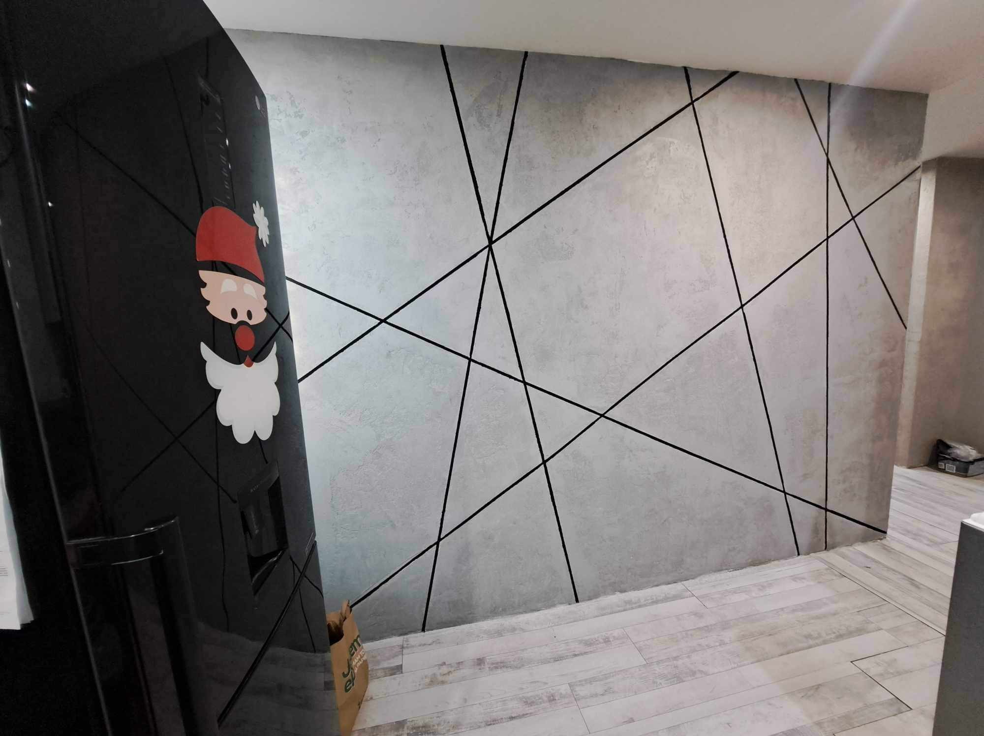 Ściana z szarą, dekoracyjną gładzią i czarnymi, geometrycznymi liniami, widziana obok czarnej lodówki z naklejką Mikołaja.