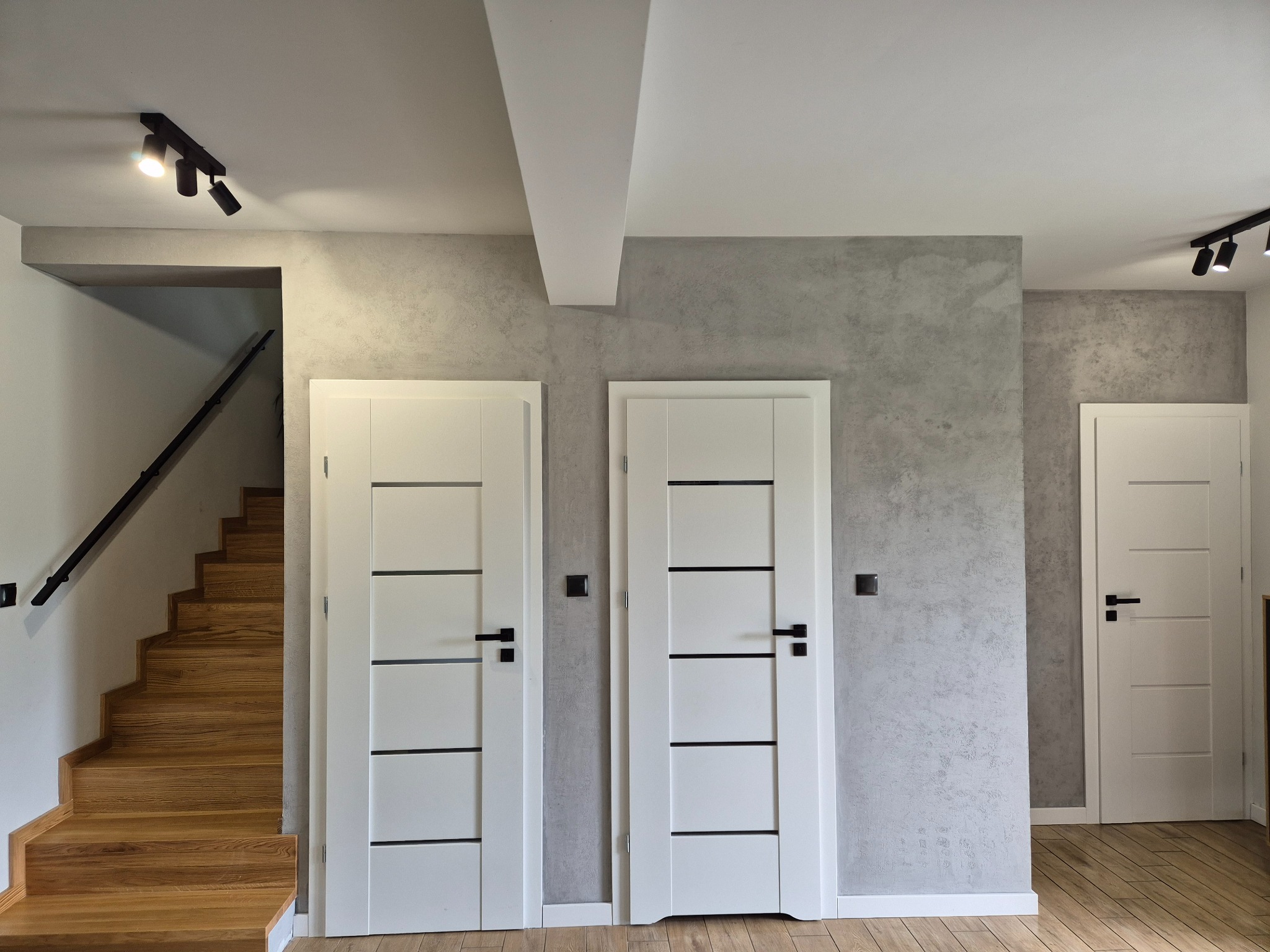 Wnętrze domu z dwiema białymi, nowoczesnymi drzwiami z czarnymi klamkami na tle ściany o szarej fakturze, drewniane schody z czarną balustradą oraz oświetlenie punktowe na suficie.