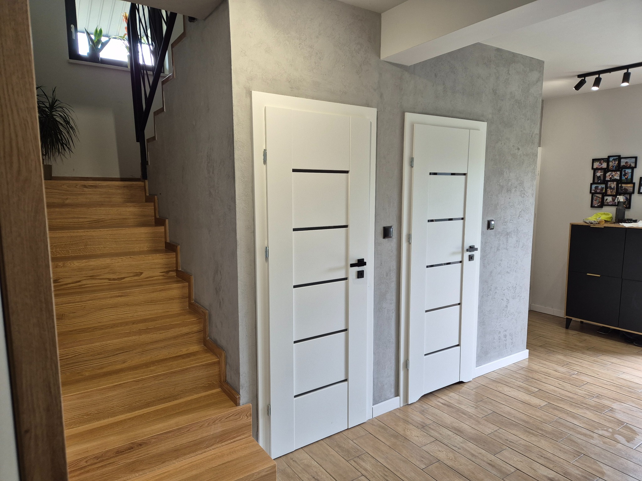 Nowoczesne wnętrze domu z drewnianymi schodami, podłogą imitującą drewno i dwiema białymi drzwiami z czarnymi akcentami na tle ściany z efektem betonu.