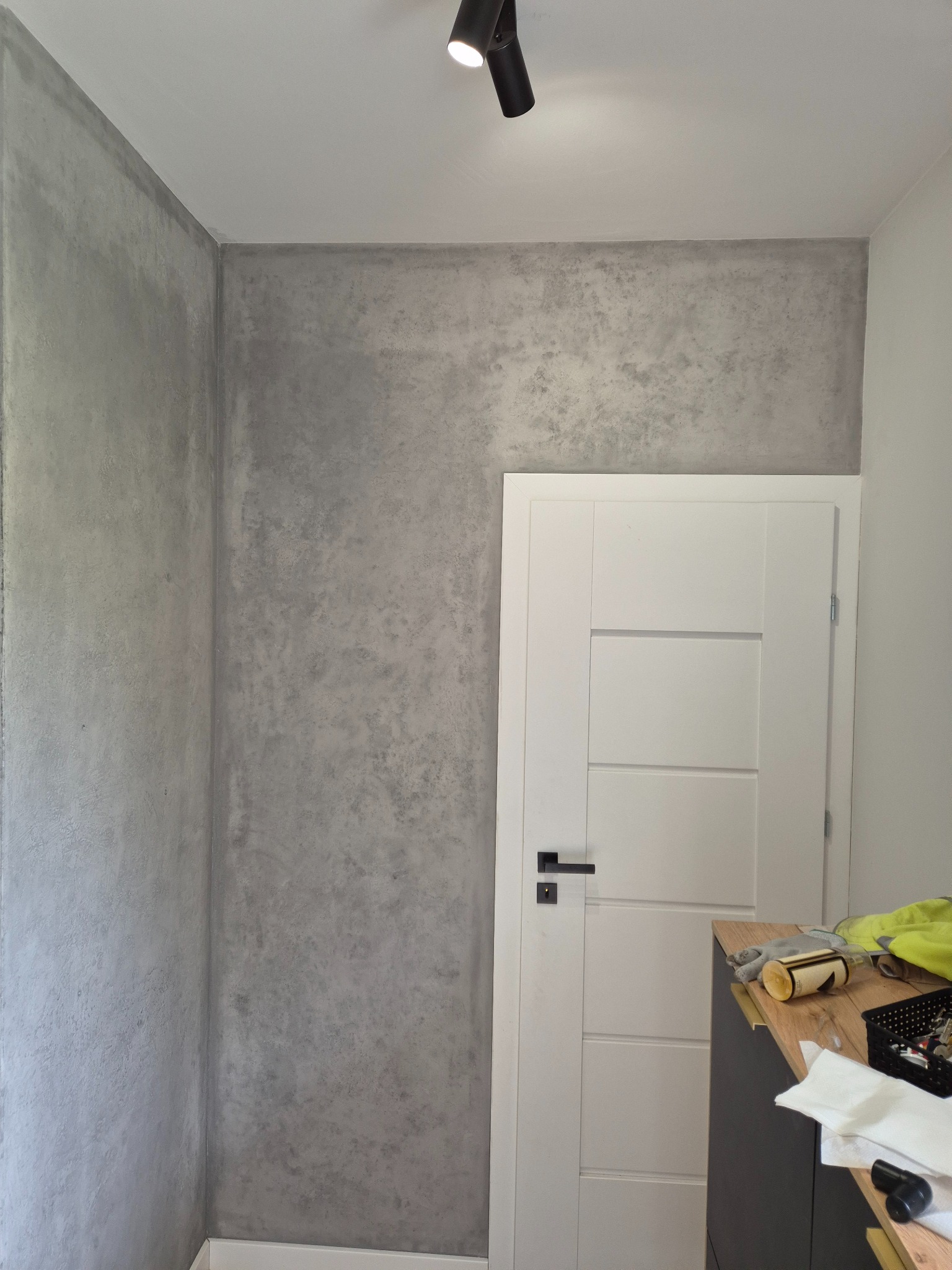 Ściana w pokoju wykończona szarą, dekoracyjną strukturą przypominającą beton, obok białe drzwi z czarną klamką i fragment szafki z narzędziami.