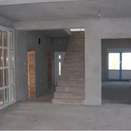 Surowe wnętrze domu w trakcie prac wykończeniowych: ściany pokryte szarym tynkiem, betonowa podłoga, widoczne schody i okna w głębi pomieszczenia.