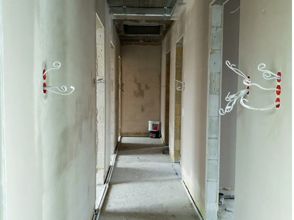 Korytarz w trakcie prac wykończeniowych: ściany po nałożeniu gładzi, wystające przewody elektryczne z puszek, widoczne niedokończone ościeżnice drzwiowe, wiadro z zaprawą w tle.