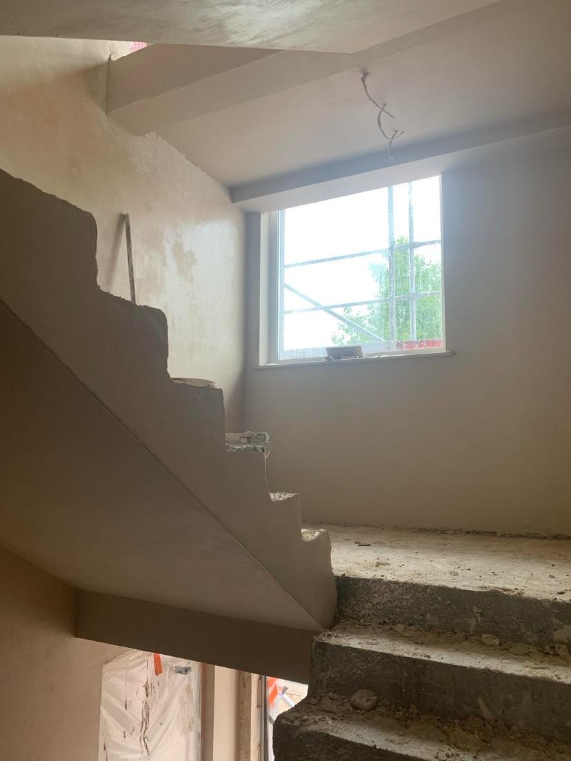 Surowe schody w trakcie prac tynkarskich, widoczne niedoskonałości i nierówności na stopniach oraz świeżo nałożony tynk na ścianach, z oknem w tle.