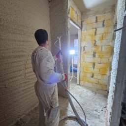 ~SERVCOMP~ EDYTA PENDZICH - Pracownik w kombinezonie, z maszyną do natrysku, nakłada świeżą warstwę tynku na ścianę w pomieszczeniu w Słupsku. Widoczna surowa ściana z pustaków i mocne oświetlenie robocze.