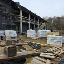 ~SERVCOMP~ EDYTA PENDZICH - Budynek w trakcie ocieplania z widocznymi rusztowaniami, izolacją termiczną na ścianach, stertami materiałów budowlanych na paletach i drewnianymi elementami szalunkowymi na pierwszym planie, przy...