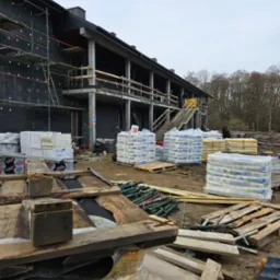 Budynek w trakcie ocieplania z widocznymi rusztowaniami, izolacją termiczną na ścianach, stertami materiałów budowlanych na paletach i drewnianymi elementami szalunkowymi na pierwszym planie, przy...