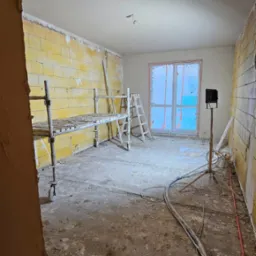 Wnętrze w trakcie prac wykończeniowych: ściany z żółtą cegłą częściowo pokryte tynkiem, rusztowanie na kółkach, drabina oparta o ścianę przy oknie, przewody na podłodze, lampa budowlana na trójnogu.