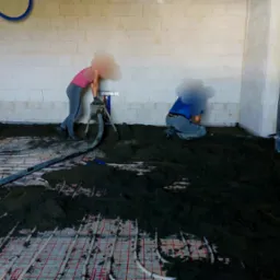 Aplikacja jastrychu na ogrzewanie podłogowe w pomieszczeniu z surowymi ścianami z betonu komórkowego; widoczne dwie osoby przy pracy i narzędzia.