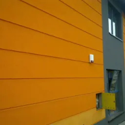 Budynek z pomarańczową elewacją panelową i szarymi wnękami okiennymi; otwarta skrzynka elektryczna z żółtymi drzwiczkami.