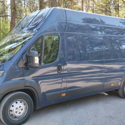 Usługi Trans Janusz Kaczorowski - Niebieski bus dostawczy z wysokim dachem zaparkowany na leśnej drodze, widok z boku, oświetlenie słoneczne.