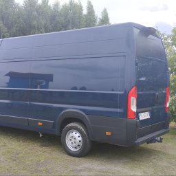 Usługi Trans Janusz Kaczorowski - Niebieski bus dostawczy zaparkowany na trawie, widok z tyłu i boku, z widoczną tablicą rejestracyjną. Tło stanowią drzewa i krzewy.