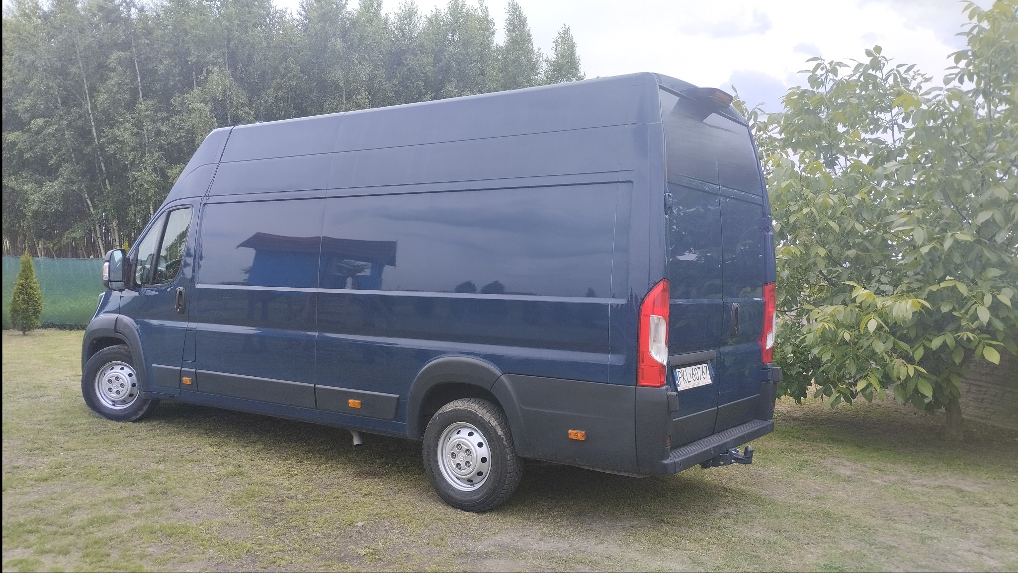 Niebieski bus dostawczy na trawie, widok z tyłu, z tablicą rejestracyjną PKL 60767. Drzewa w tle. Pojazd gotowy do transportu.