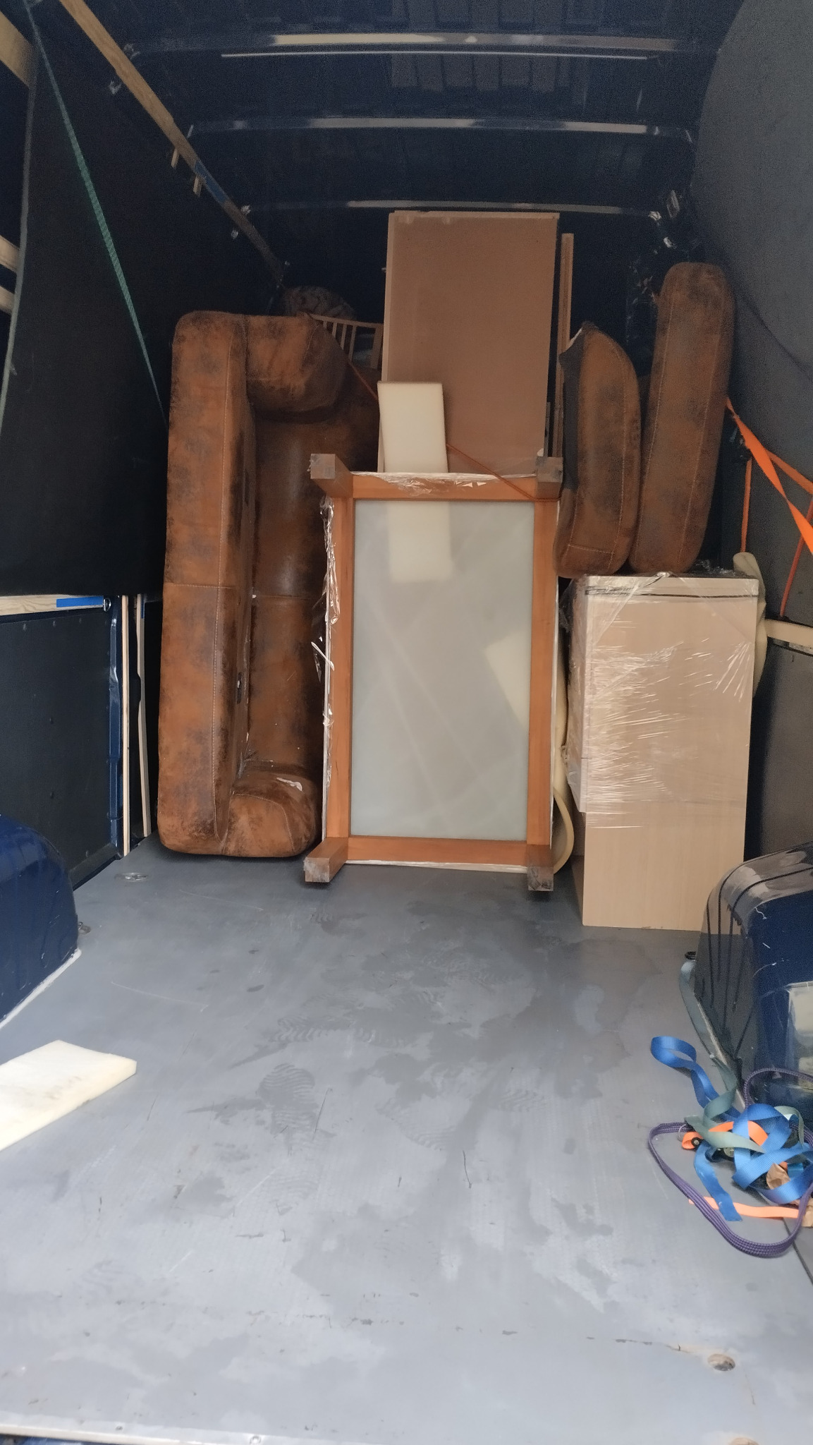 Wnętrze busa wypełnione meblami: sofa, lustro w ramie, zabezpieczone folią i pasami. Widok z tyłu pojazdu. Ślady stóp na podłodze. Nierównomierne oświetlenie.