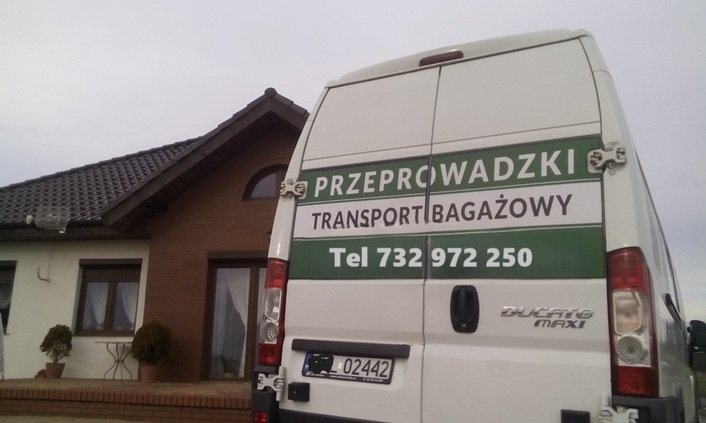 Tył białego busa Ducato Maxi z zielonym napisem 'Przeprowadzki Transport Bagażowy' i numerem telefonu, zaparkowanego przed domem.