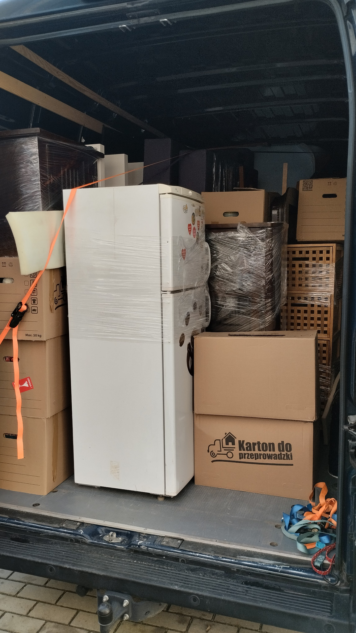 Wnętrze busa wypełnione kartonami, meblami i białą lodówką zabezpieczoną folią stretch, gotowe do transportu. Widoczne pasy mocujące i kartony z napisem 'Karton do przeprowadzki'.