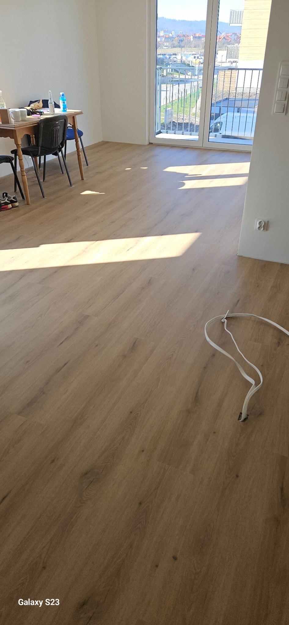 Jasny, nowo położony panel podłogowy w pustym pokoju z widokiem na miasto przez balkon, z przewodami elektrycznymi leżącymi na podłodze w prawym dolnym rogu.