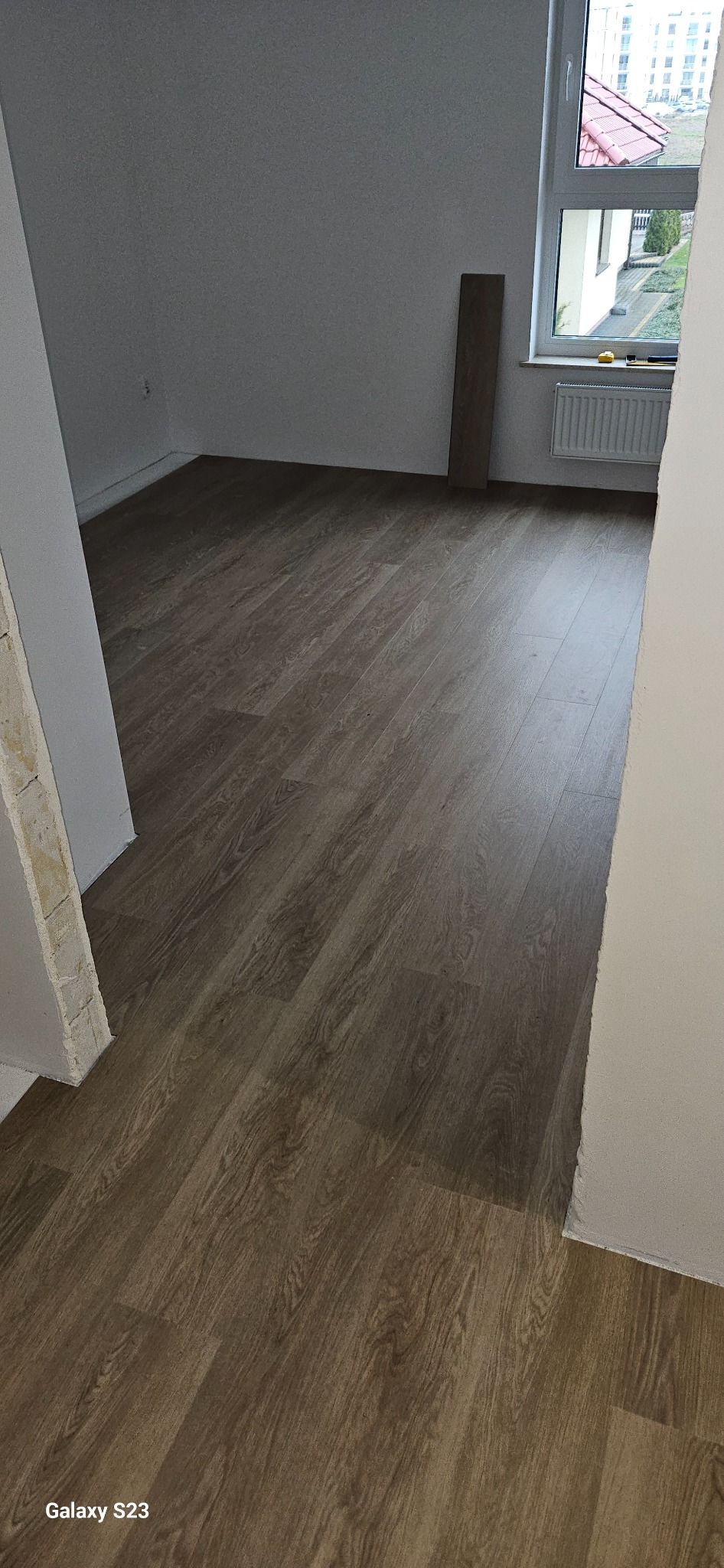 Ułożone panele podłogowe w pokoju, widok z progu, widoczny fragment ściany z oknem i grzejnikiem oraz niedokończone obramowanie drzwi.