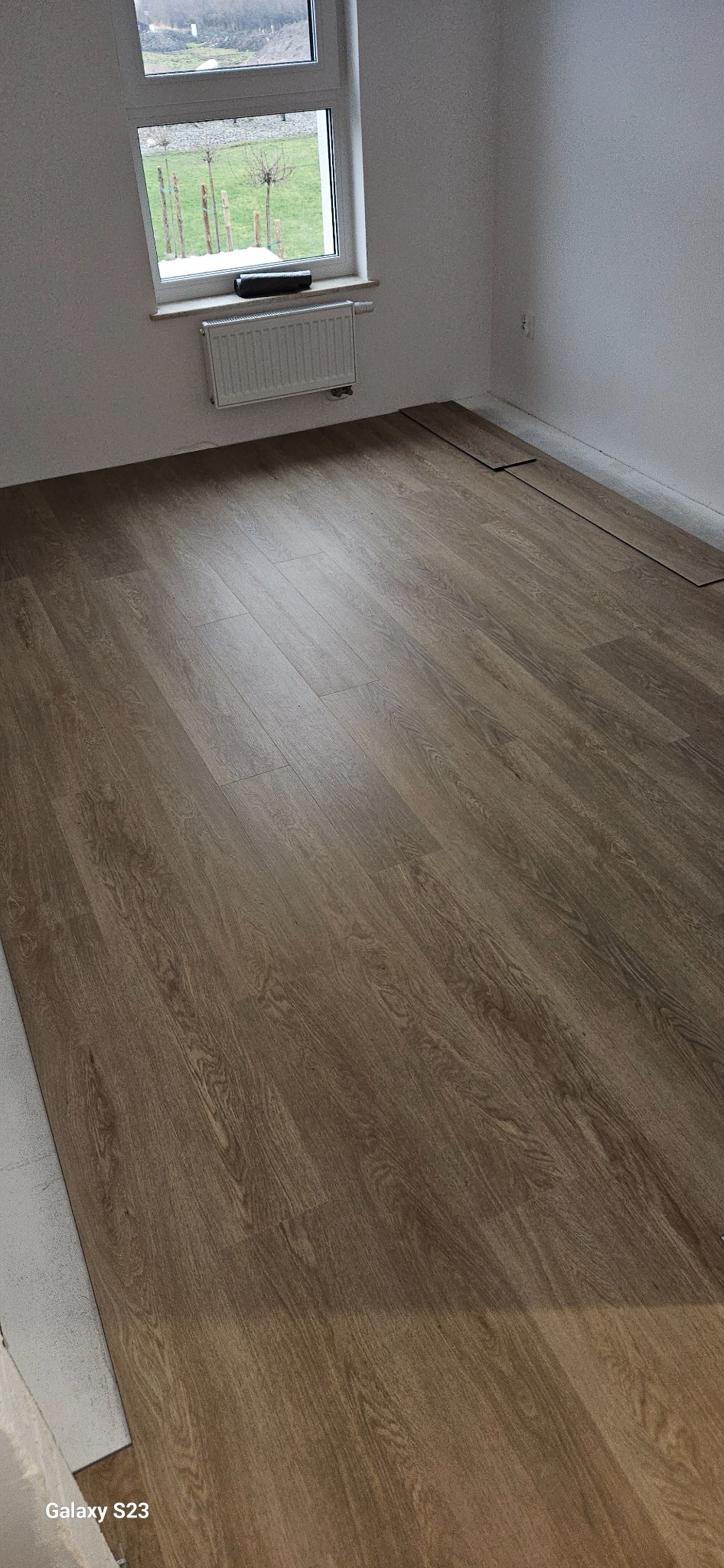 Fragment pokoju z ułożonymi panelami podłogowymi w odcieniach brązu, widoczne niedokończone fragmenty przy ścianie i grzejniku, za oknem widok na ogród zimą.