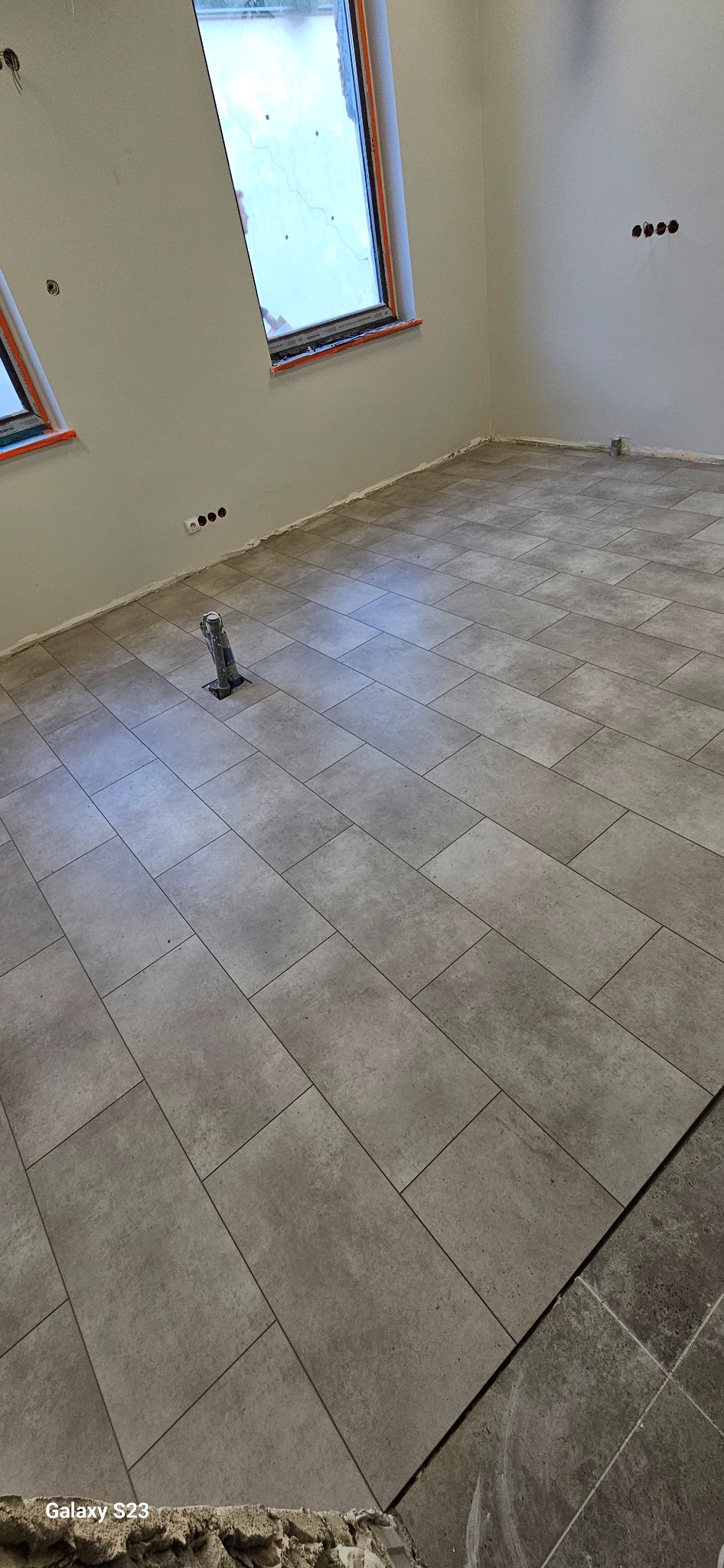 Ułożona podłoga z szarych, prostokątnych płytek w nowym pomieszczeniu z oknami dachowymi i wystającymi rurami instalacji. Fragment starej podłogi widoczny w rogu.