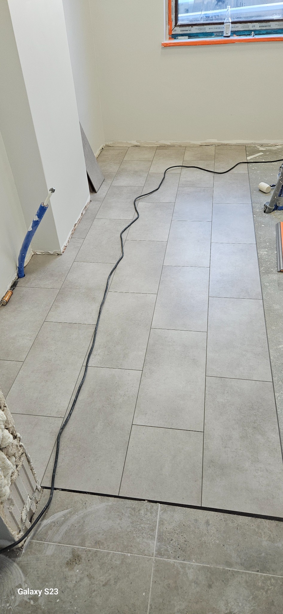 Układanie płytek podłogowych w jasnym odcieniu szarości, widoczny kabel zasilający narzędzie, fragment nieukończonej ściany z pianką montażową oraz okno z pomarańczowymi uszczelkami.