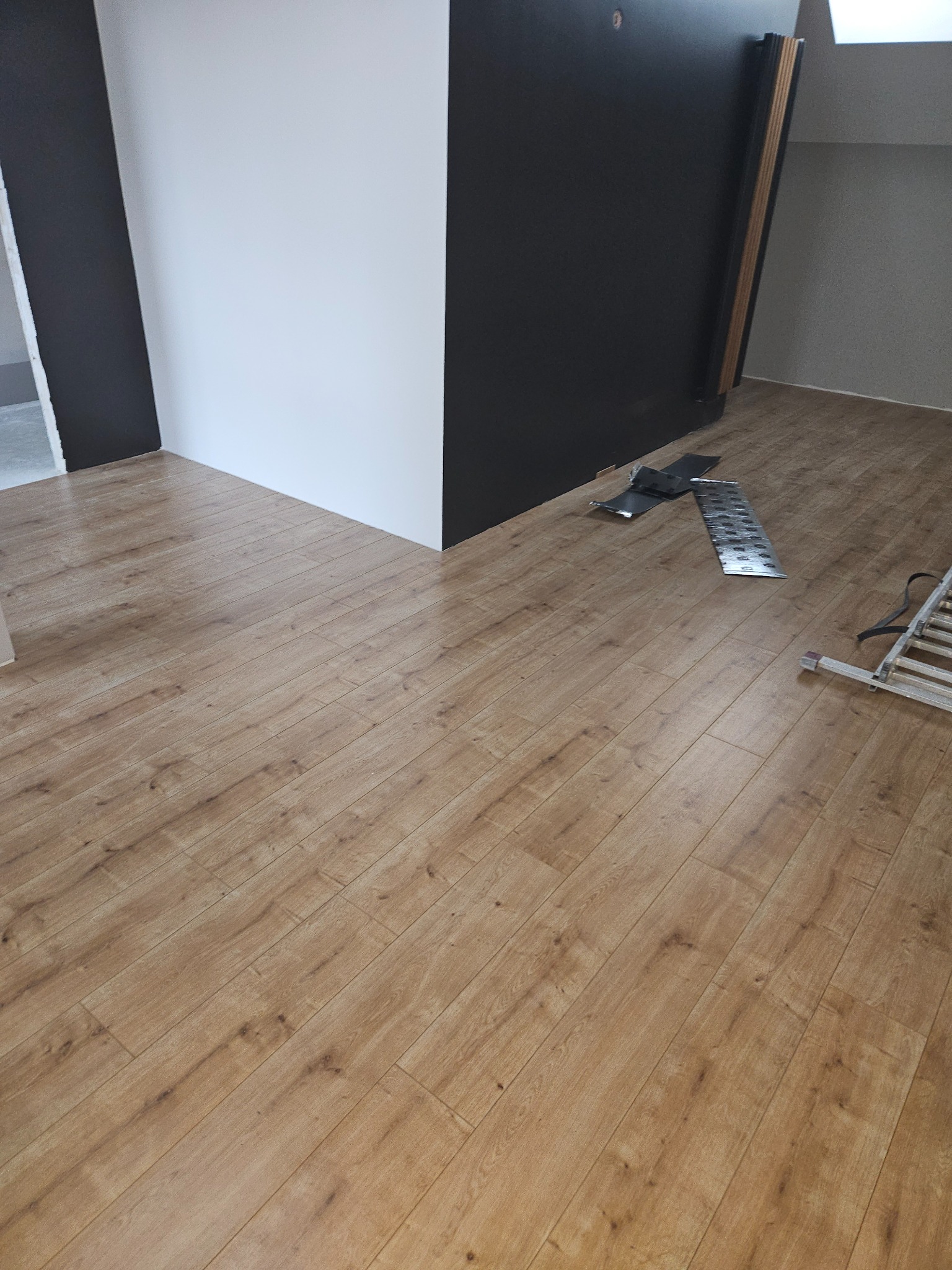 Ułożone panele podłogowe w odcieniach brązu w pokoju z białymi i czarnymi ścianami, widoczne akcesoria montażowe i fragment drabiny.