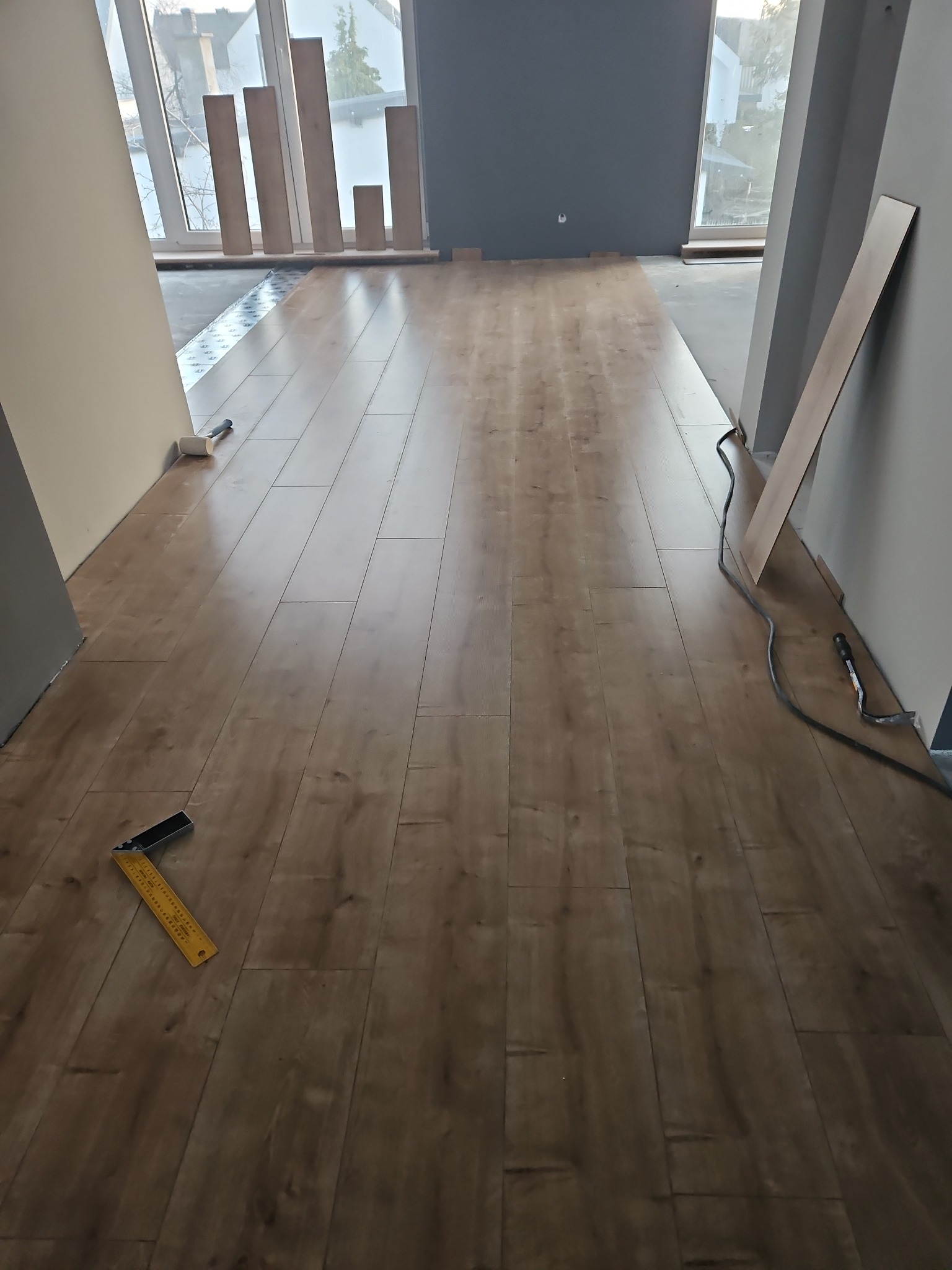 Częściowo ułożona podłoga z paneli w jasnym odcieniu drewna, widoczny kątownik stolarski, młotek gumowy i nieużyte panele pod oknem. Na podłodze widoczne ślady lekkiego zabrudzenia.